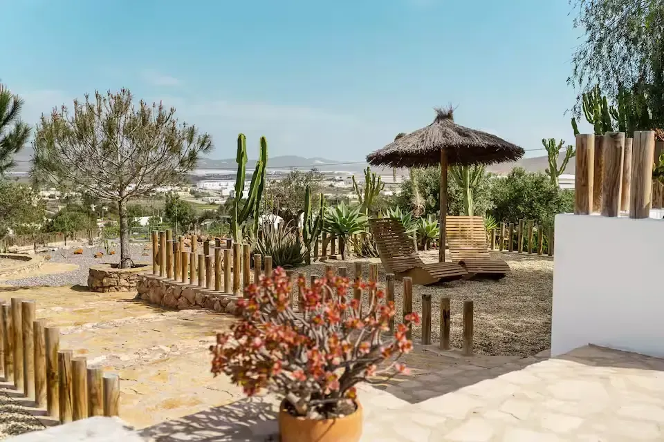 Patio soleado con jardín, macetas con flores, sombrilla de paja y valla de madera en un paisaje desértico.