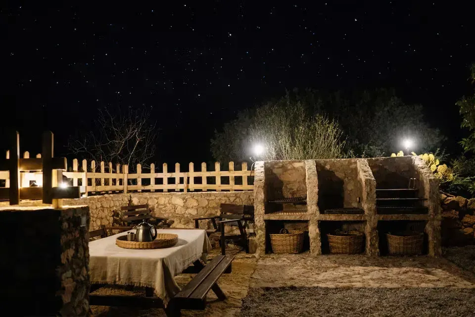 Patio exterior por la noche con mesa, sillas, parrilla de piedra y guirnaldas de luces bajo un cielo estrellado.