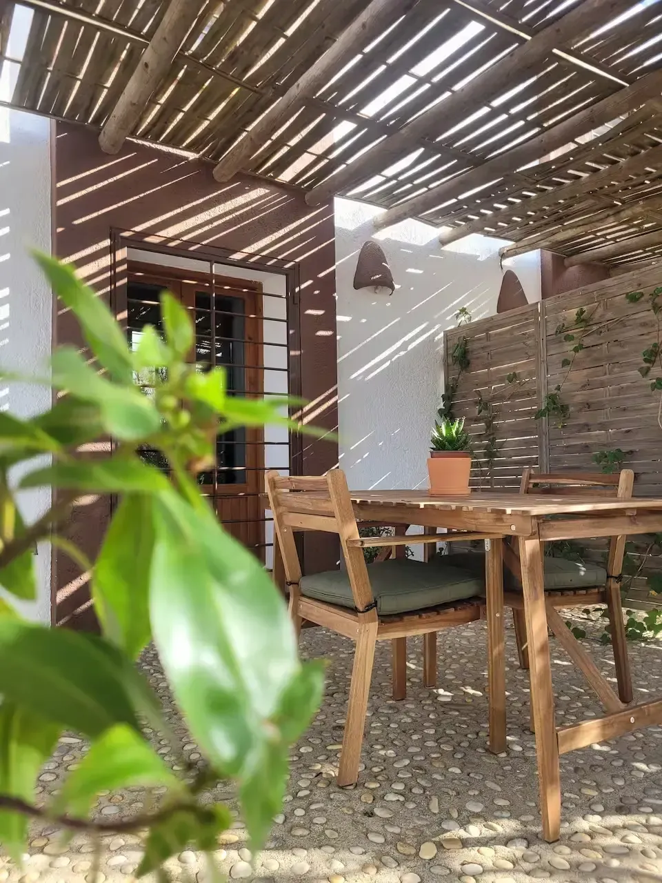 Patio exterior con mesa y sillas de madera bajo un techo de listones, luz solar y una planta verde en primer plano.