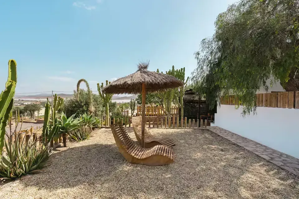 Patio bohemio con tumbonas de mimbre, sombrilla de paja, suelo de grava y plantas del desierto bajo un cielo azul.