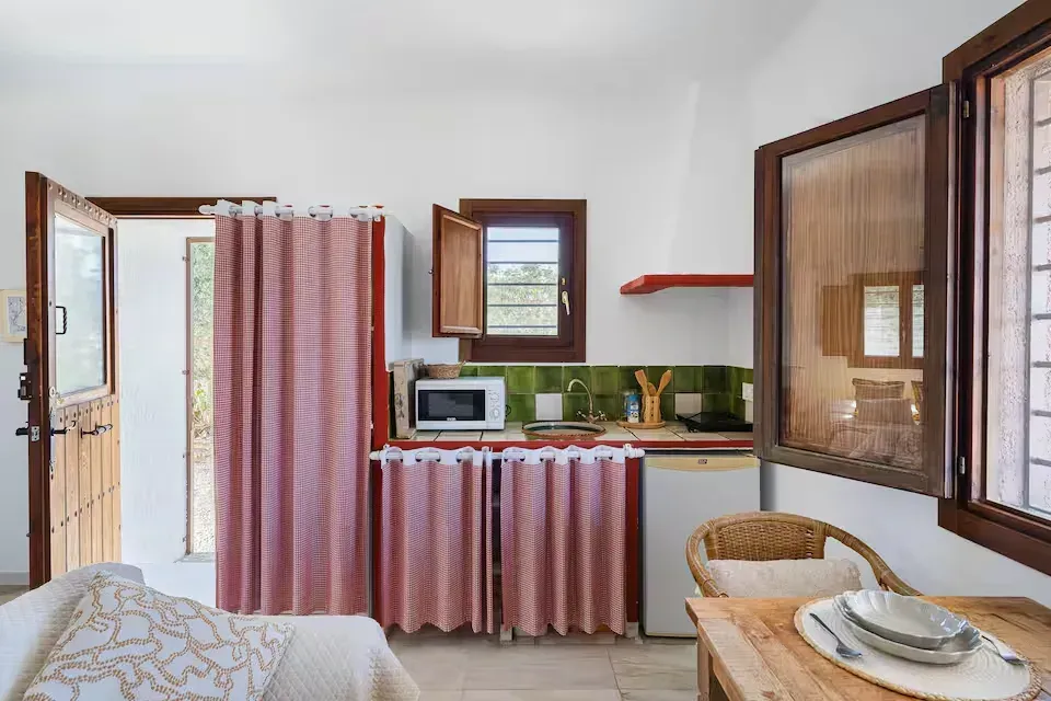 Cocina pequeña con cortinas rosas, microondas, silla de mimbre y contraventanas de madera abiertas.