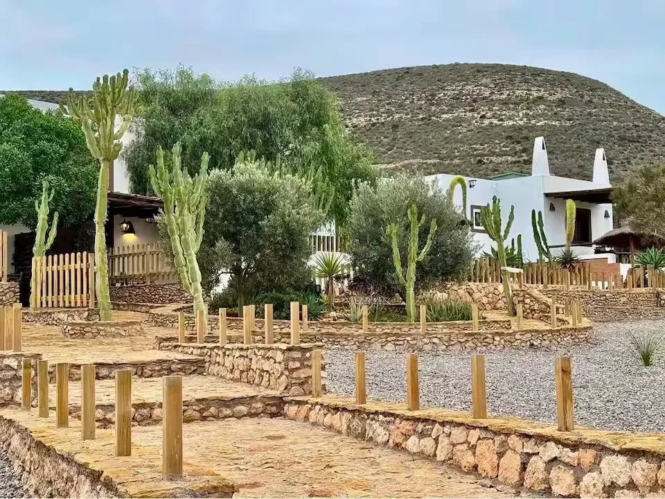 Casas blancas rurales junto a un sendero rocoso y vallas de madera, con árboles frondosos y una colina al fondo.