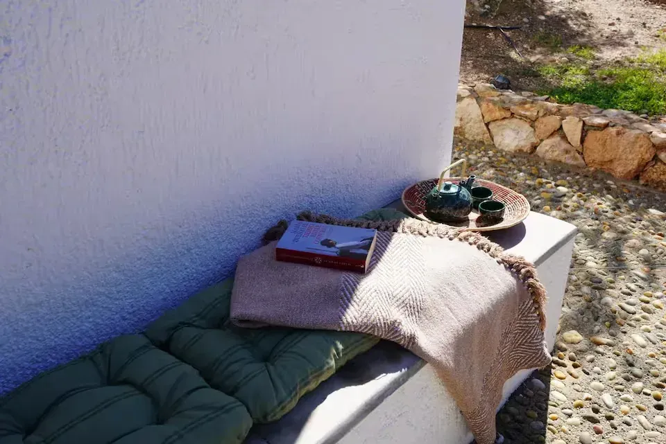 Un banco acolchado con manta y bandejas junto a una pared blanca en un patio soleado al aire libre.