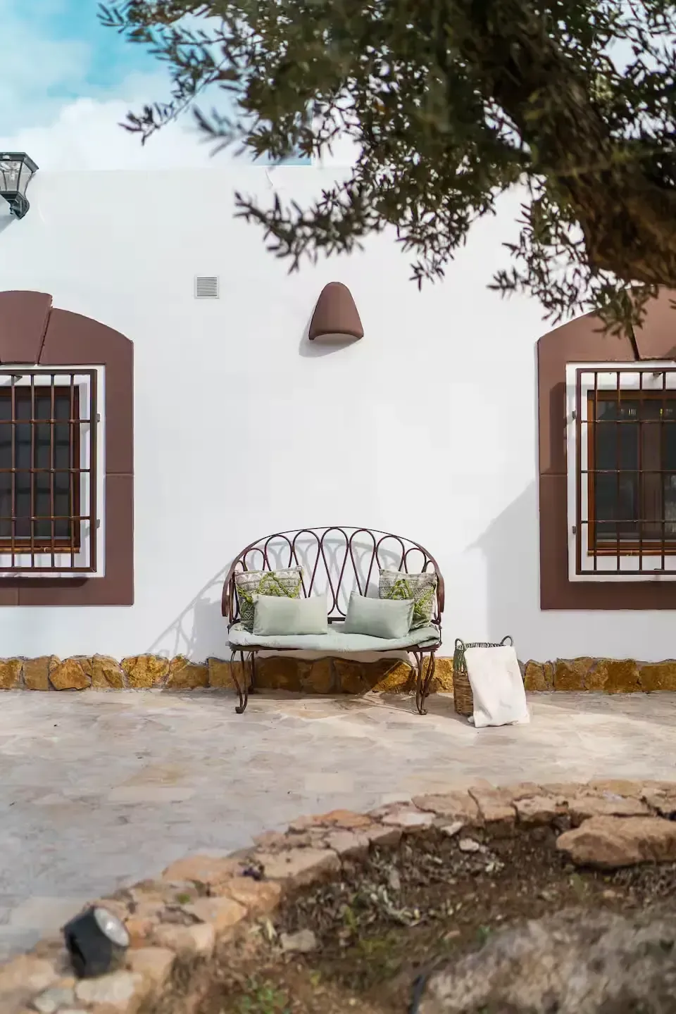 Fachada de una casa de estuco blanco con dos ventanas enrejadas y un banco de metal bajo un árbol