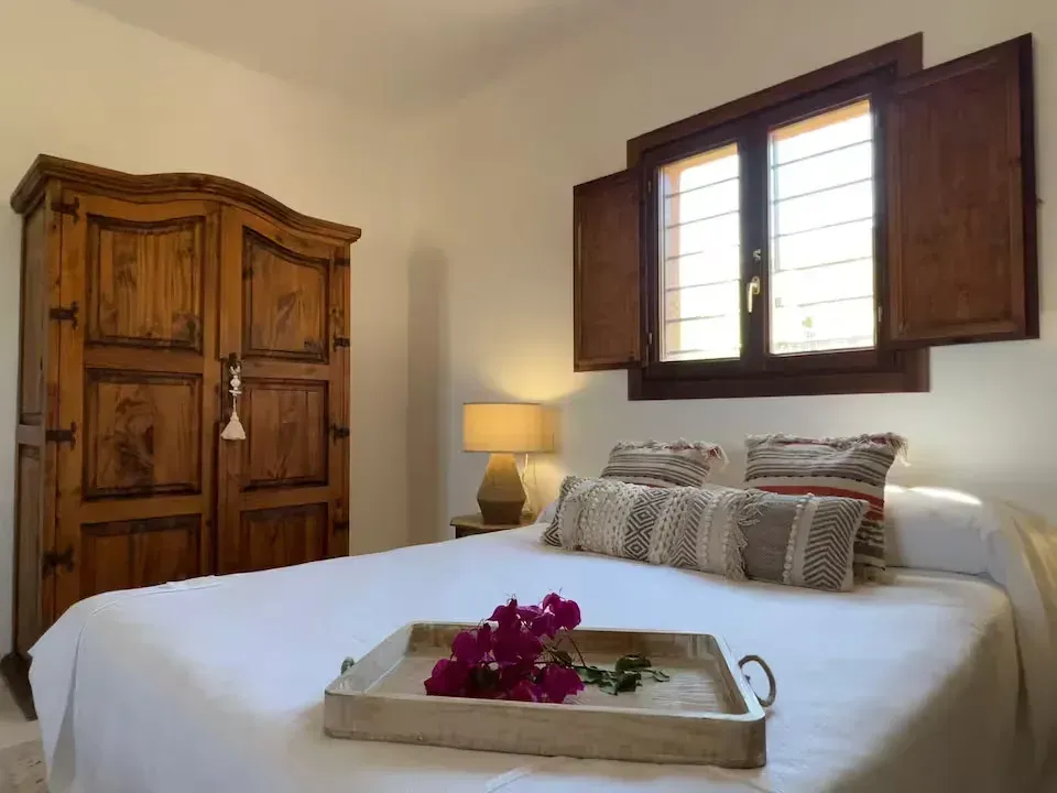 Dormitorio con cama blanca, armario de madera, ventana abierta y bandeja con flores moradas sobre la cama.