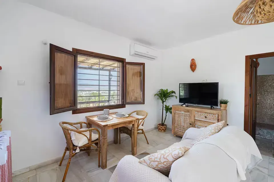 Luminoso salón con juego de comedor de mimbre, sofá blanco, mueble para la televisión y contraventanas abiertas.
