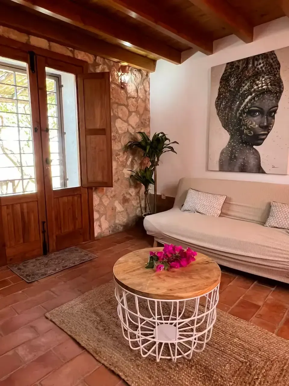 Acogedora sala de estar rústica con vigas de madera, pared de piedra, sofá beige, mesa de centro redonda y flores rosas.