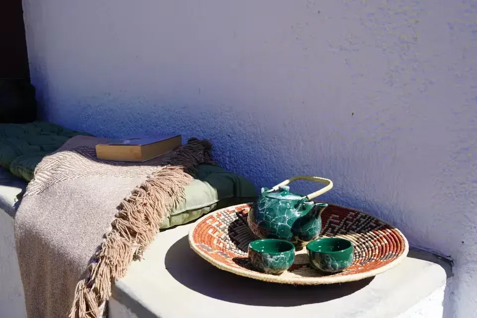 Un juego de té sobre una bandeja encima de una mesa blanca junto a un banco con una manta doblada contra una pared azul.