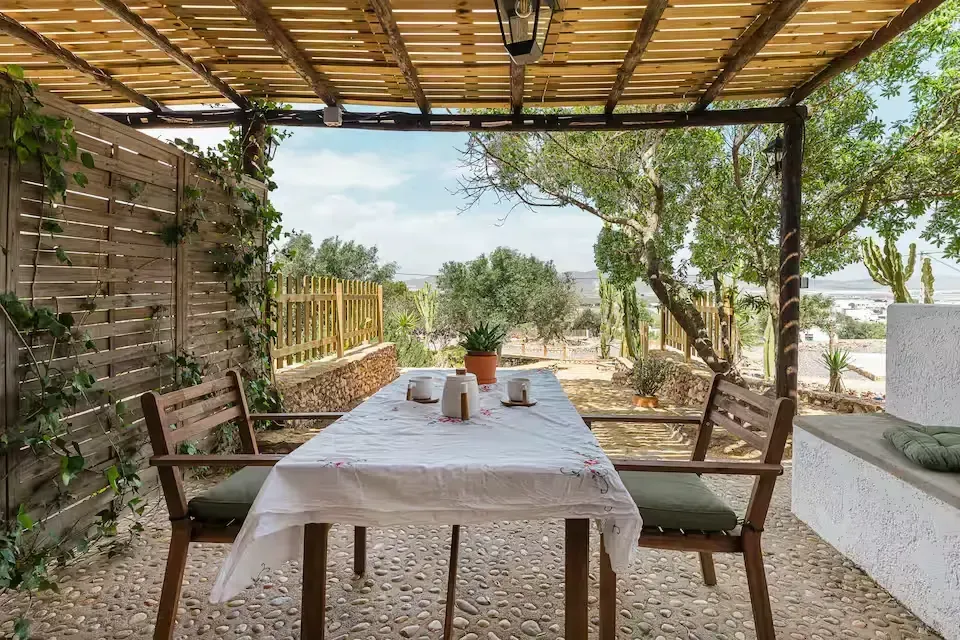 Mesa de comedor en el patio cubierto con mantel blanco, sillas y vista al jardín iluminado por el sol.