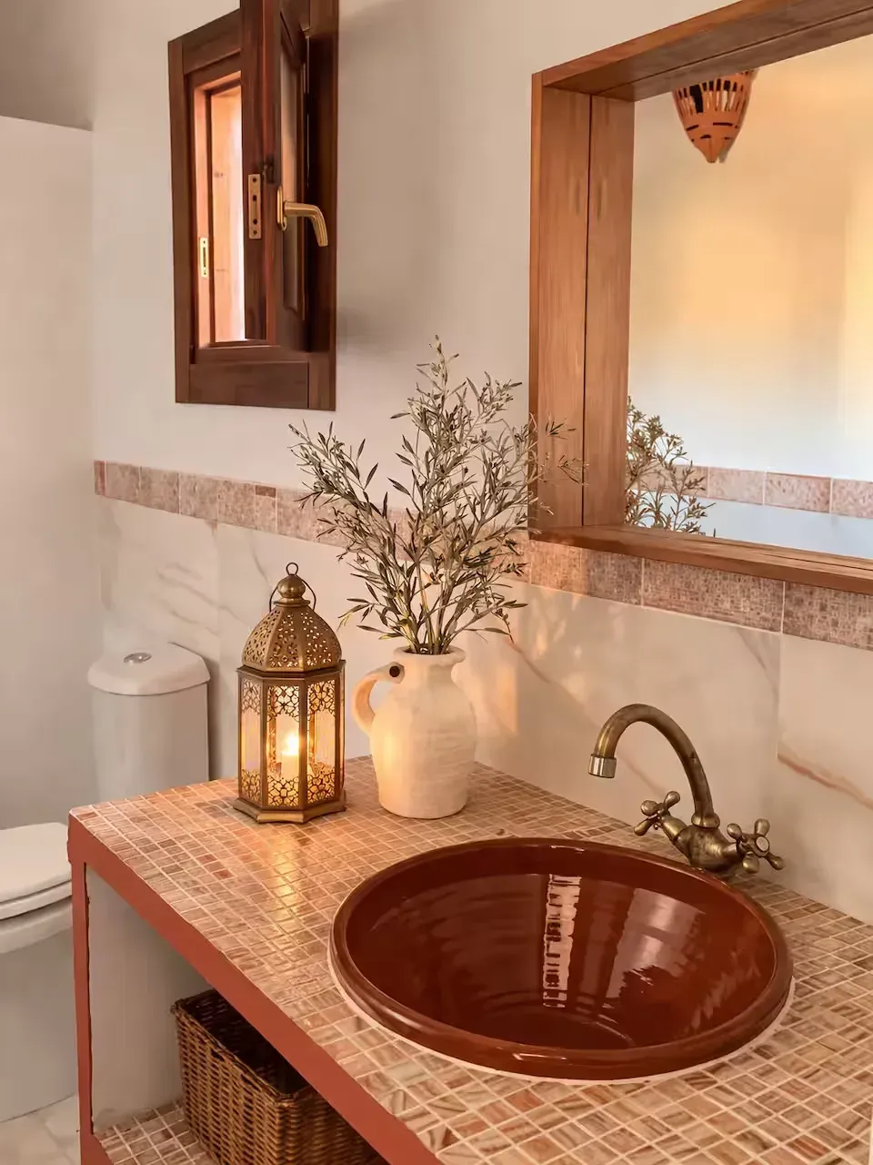 Tocador de baño rústico con lavabo de cobre, espejo, farol y jarrón blanco sobre una encimera de madera.
