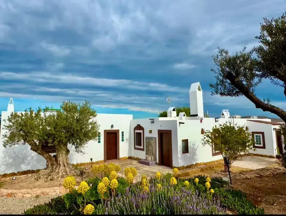 Casa blanca con detalles azules y flores de jardín bajo un cielo nublado, enmarcada por árboles y un camino de piedra.