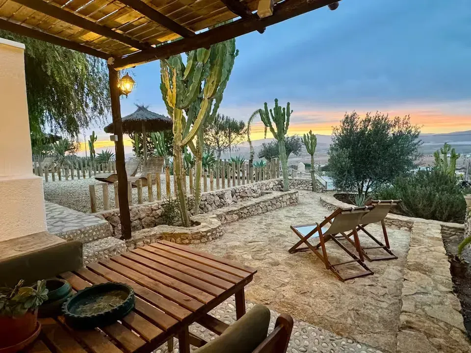 Patio de piedra con mesa y sillas de madera, cactus y vista al atardecer sobre el agua.