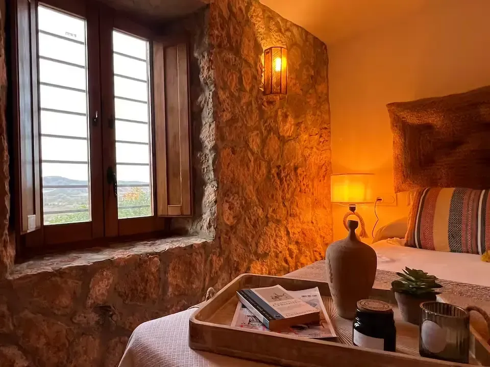 Acogedora habitación rústica con paredes de piedra, vistas a la ventana, luz cálida de lámpara y una bandeja sobre la cama.