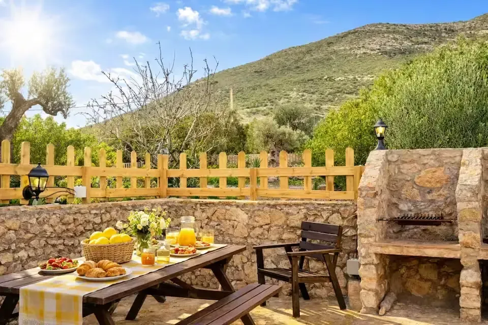 Patio exterior con mesa de madera, fruta, parrilla de piedra y vistas a la montaña tras una valla amarilla.