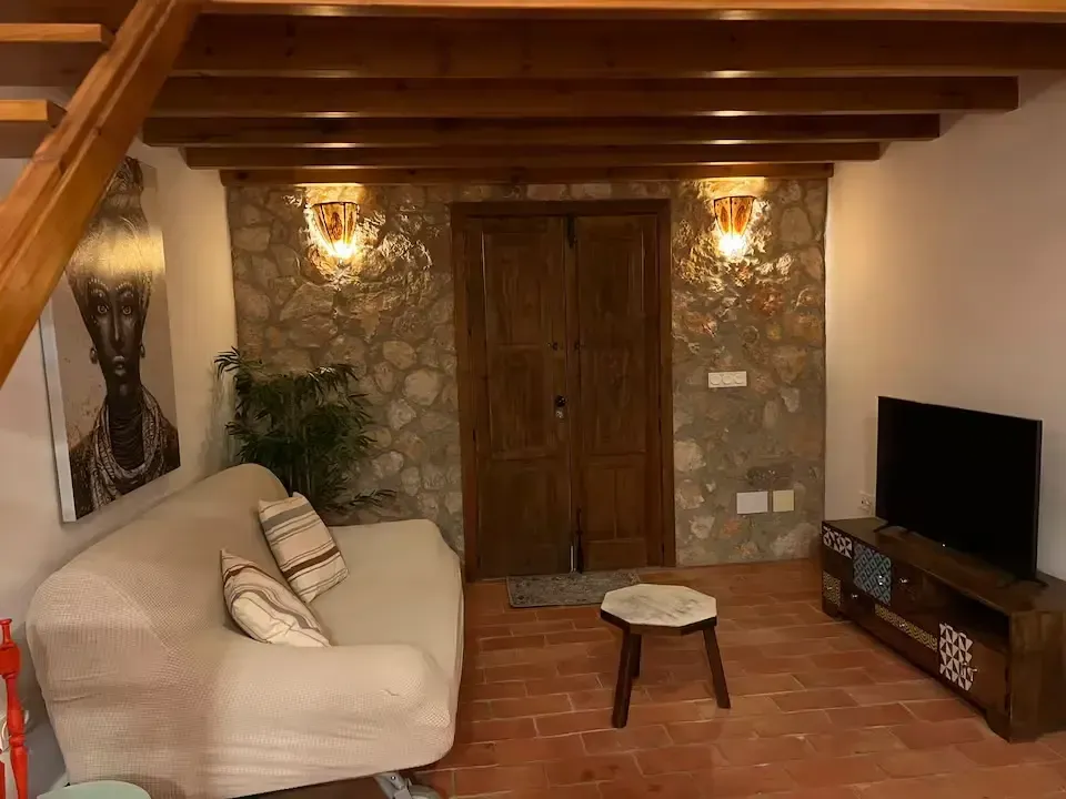 Acogedora sala de estar con sofá beige, pared de piedra, vigas de madera en el techo y televisor sobre un soporte.