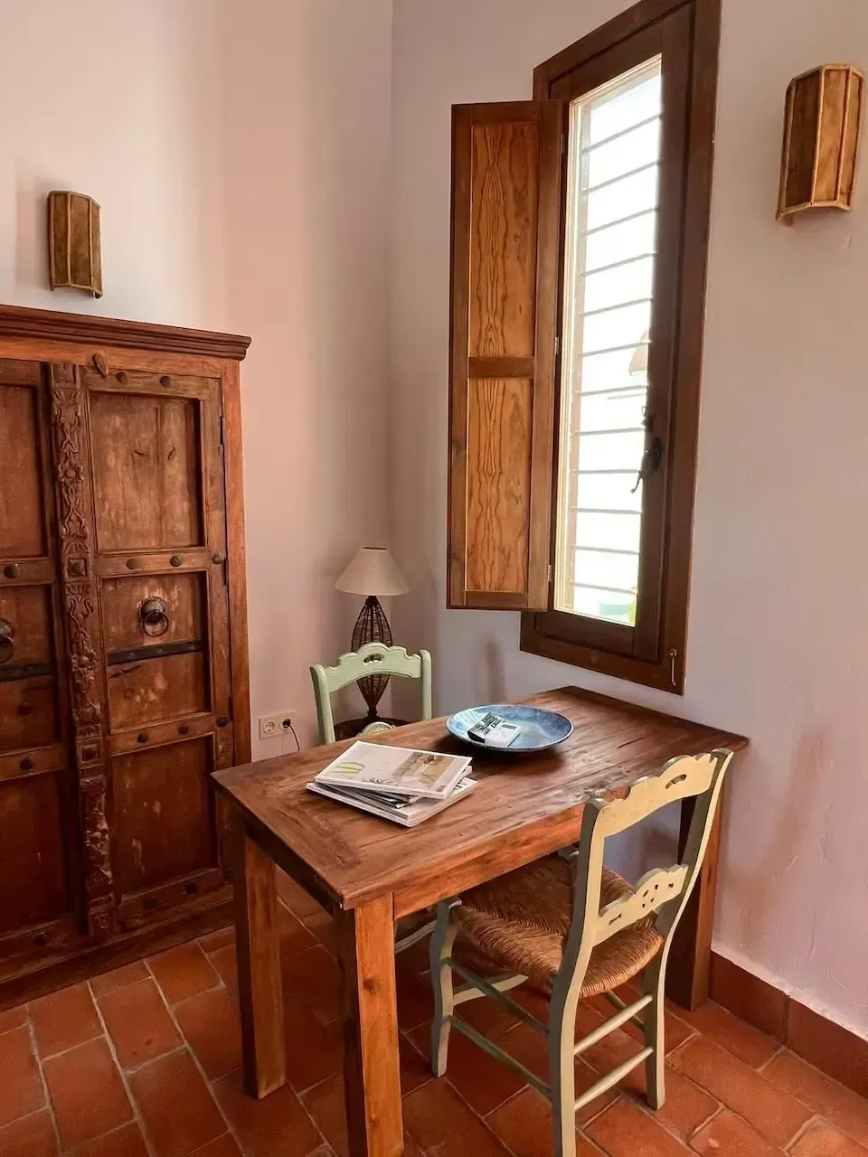 Pequeña habitación rústica con escritorio de madera, silla, armario y ventana que deja entrar la luz.