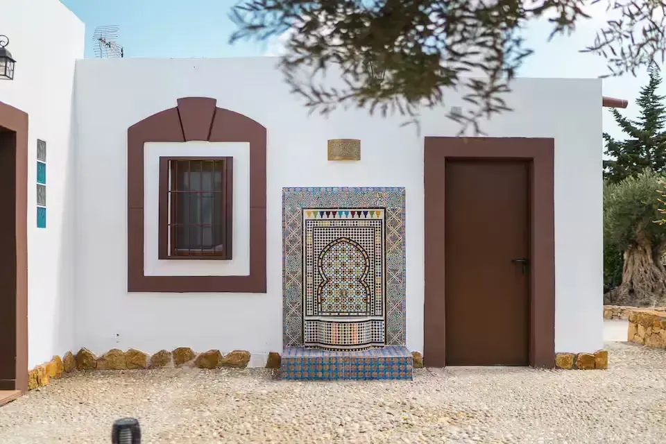 Fachada de casa de estuco blanco con ventanas con marcos marrones, una fuente decorativa de azulejos y una puerta marrón.