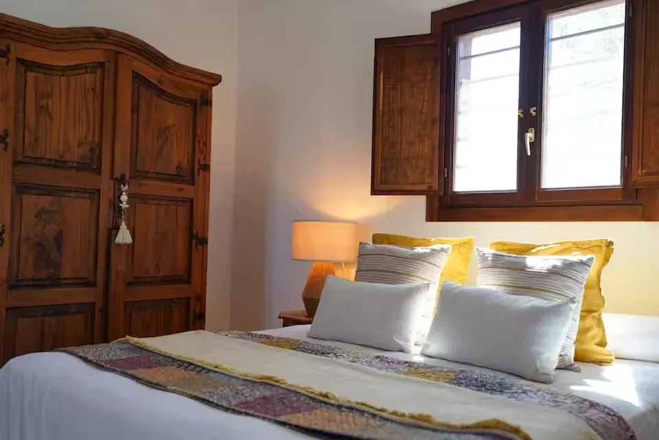 Acogedora habitación con armario de madera, cama con ropa de cama blanca y amarilla, lámpara y contraventanas.