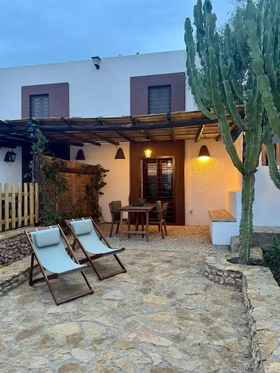 Patio de piedra con tumbonas, mesa de comedor y luces cálidas junto a una casa blanca y un cactus alto al atardecer.