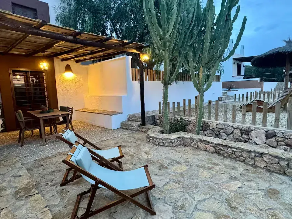 Patio de piedra con tumbonas junto a una zona de comedor exterior iluminada y cactus al atardecer.