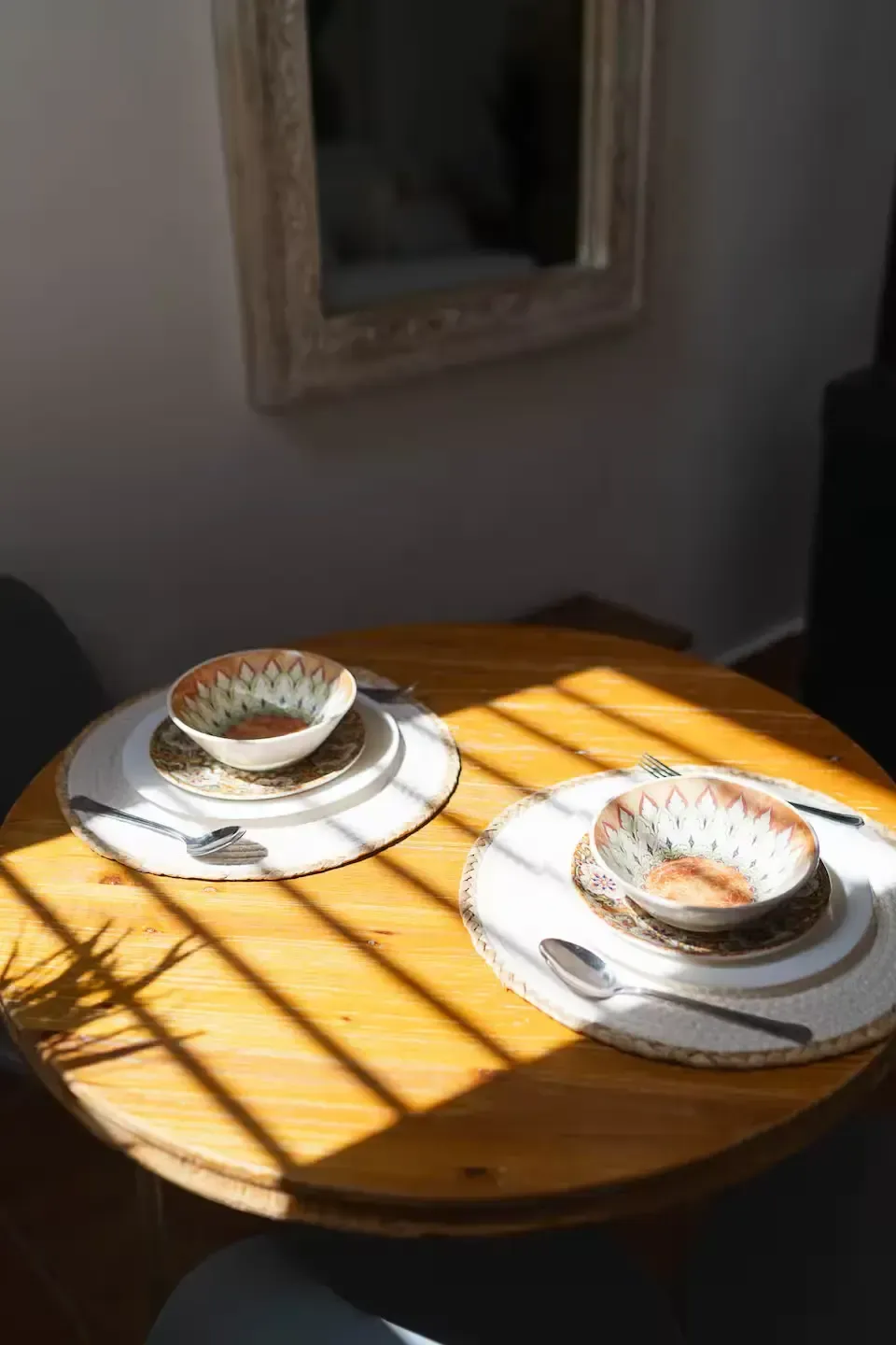 Mesa redonda de madera iluminada por el sol con dos tazas de café y sus platillos cerca de una ventana.