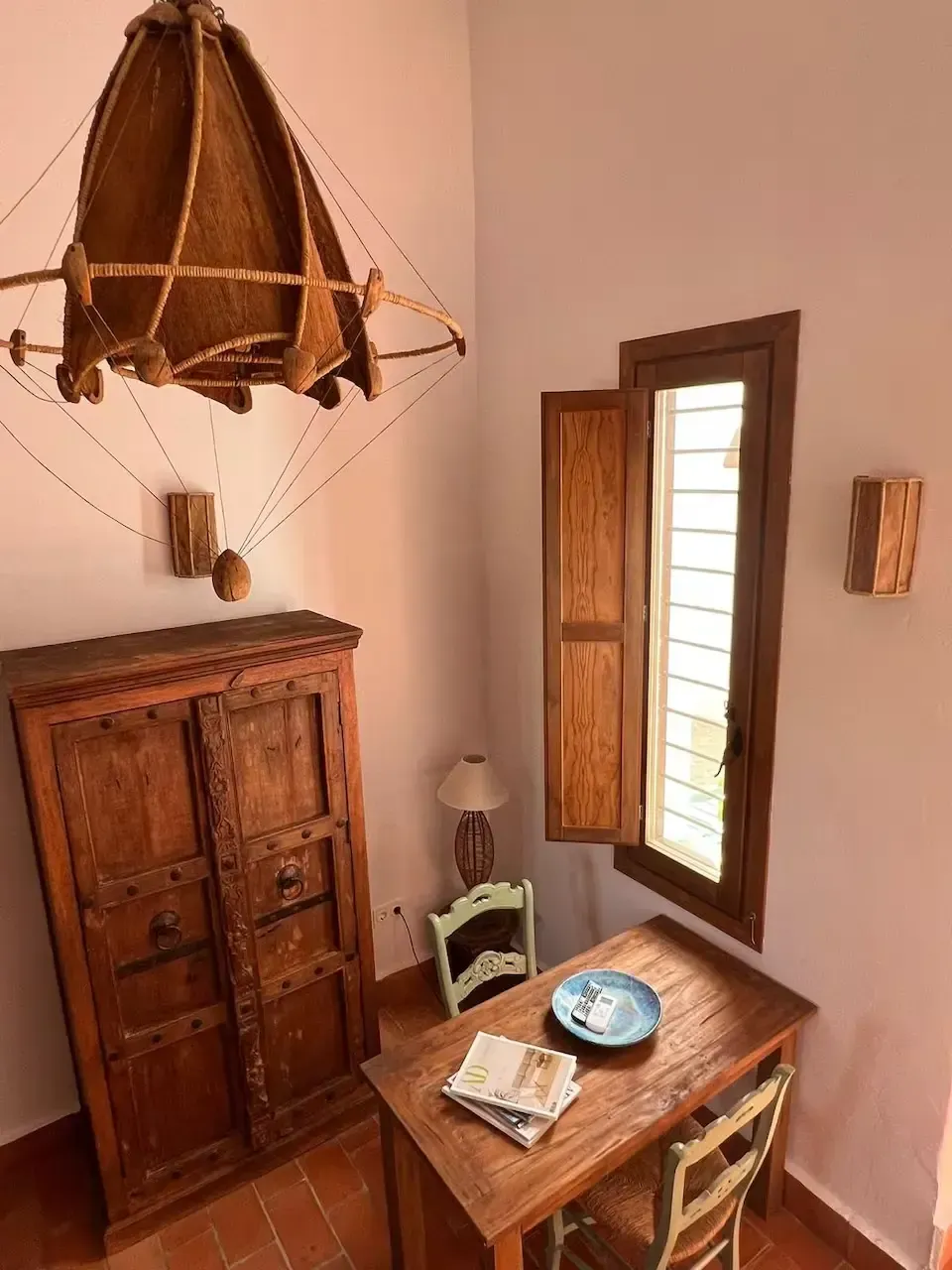 Pequeña habitación rústica con armario de madera, escritorio, ventana abierta y lámpara colgante de mimbre.