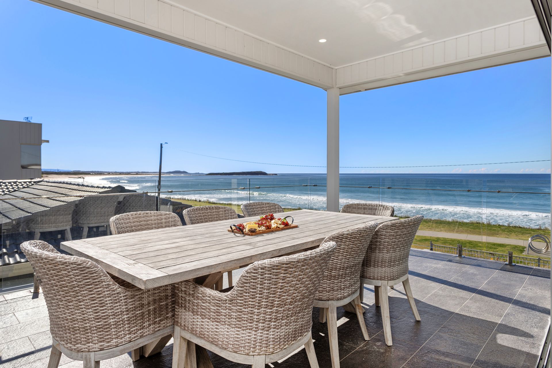 Outdoor dining area overlooking the ocean. White table and woven chairs on a tiled deck with blue sky.