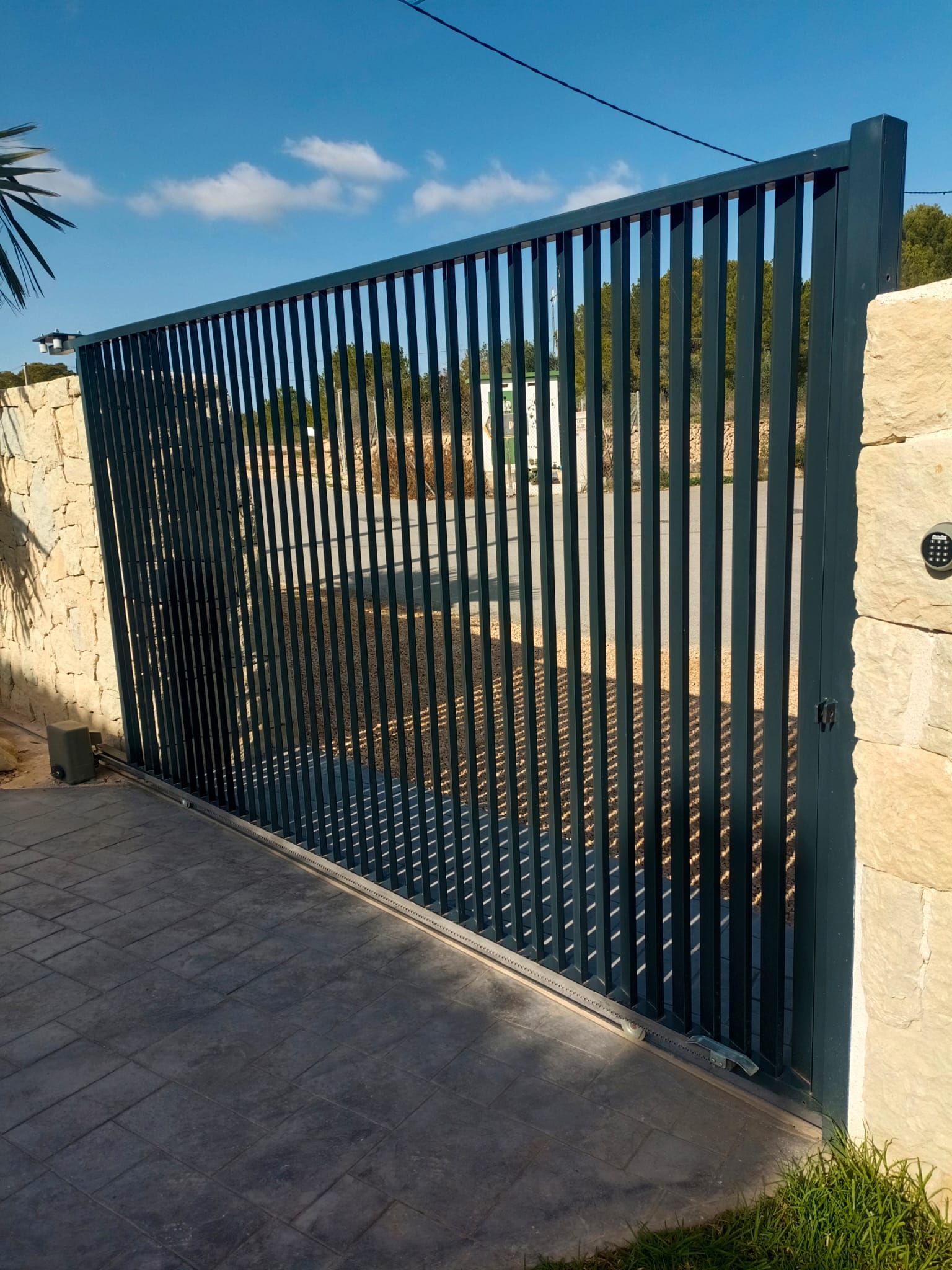 Puerta de lamas metálicas de color gris situada entre muros de piedra sobre una superficie de hormigón.