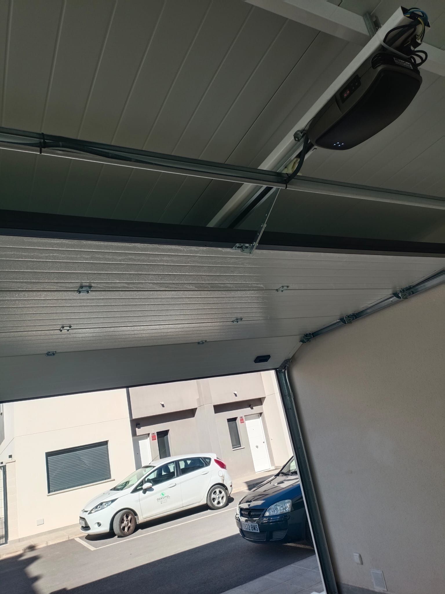 La puerta del garaje está parcialmente abierta, dejando ver un coche blanco aparcado en el exterior. Se ven un motor y un sistema de raíles.