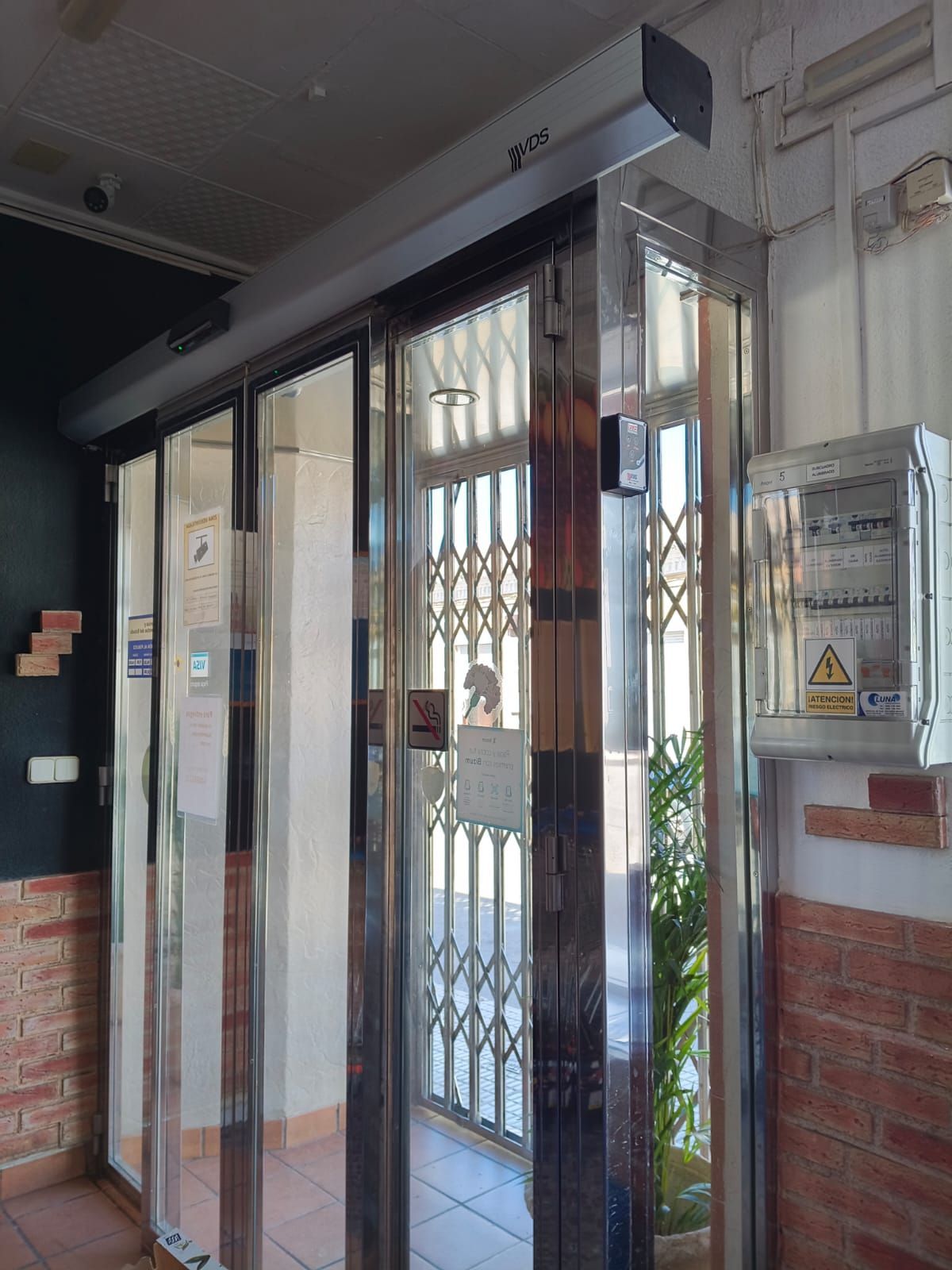 Ascensor con puertas de cristal y estructura metálica, dentro de un edificio. Puerta de seguridad visible. Cuadro eléctrico a la derecha.