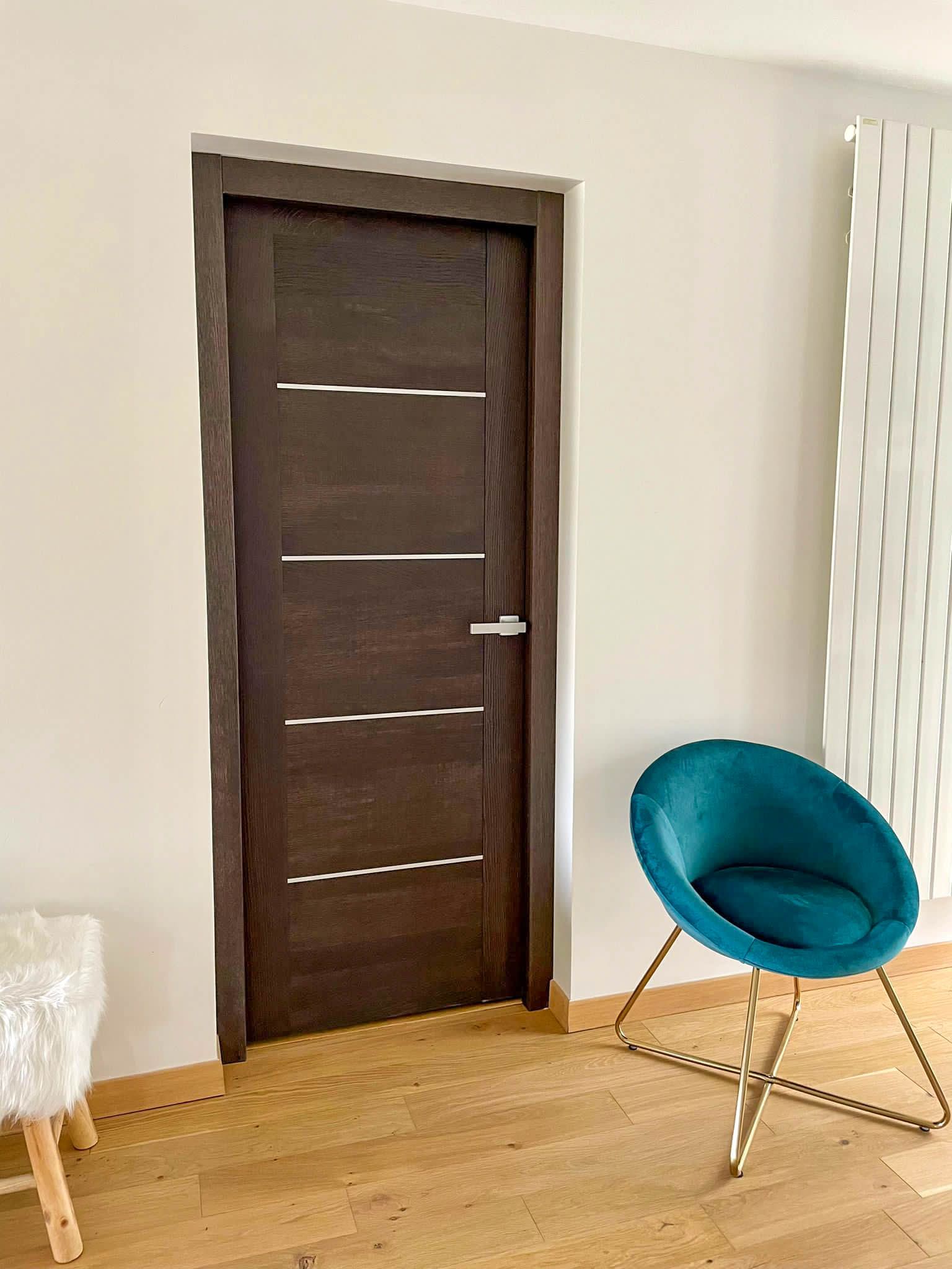 Espace intérieur moderne avec porte en bois foncé ornée de bandes métalliques horizontales. À droite, un fauteuil bleu canard aux pieds dorés. Sol en bois clair et tabouret blanc en fourrure visible à gauche.