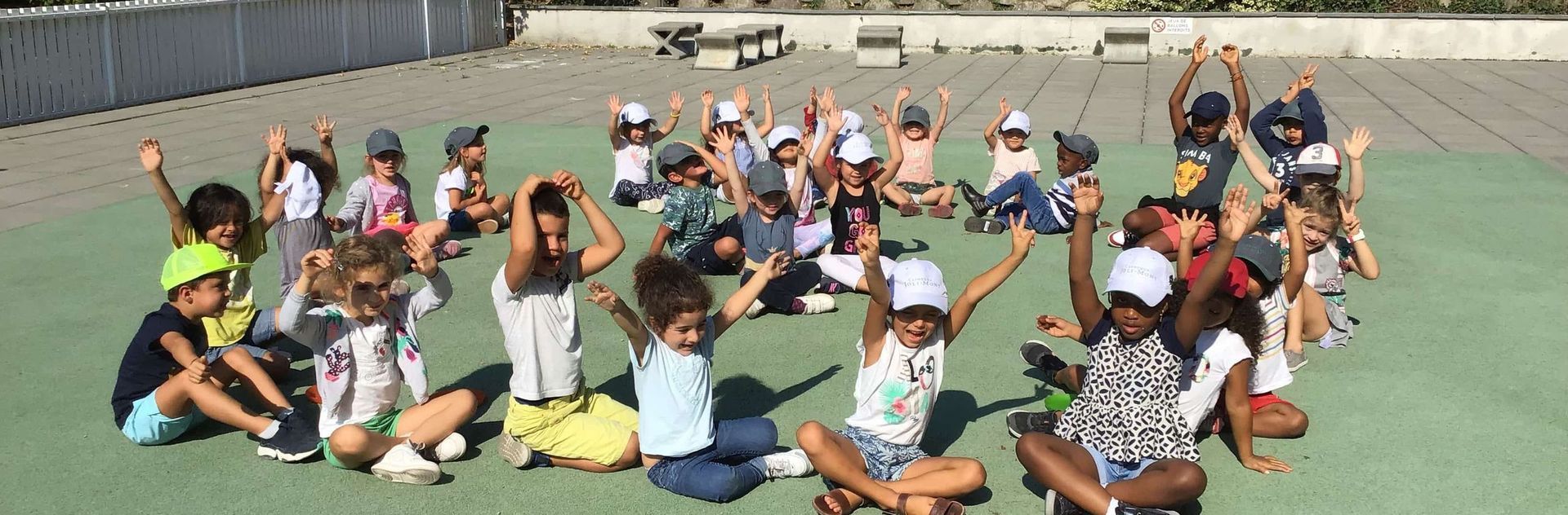 Groupe d'enfants assis en cercle, les bras levés, sur une surface verte.