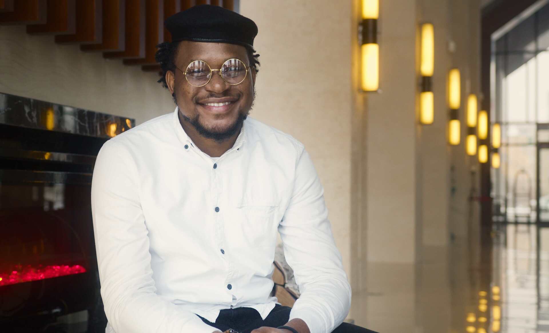 Ein Mann mit Brille und Hut sitzt auf einem Hocker und lächelt.