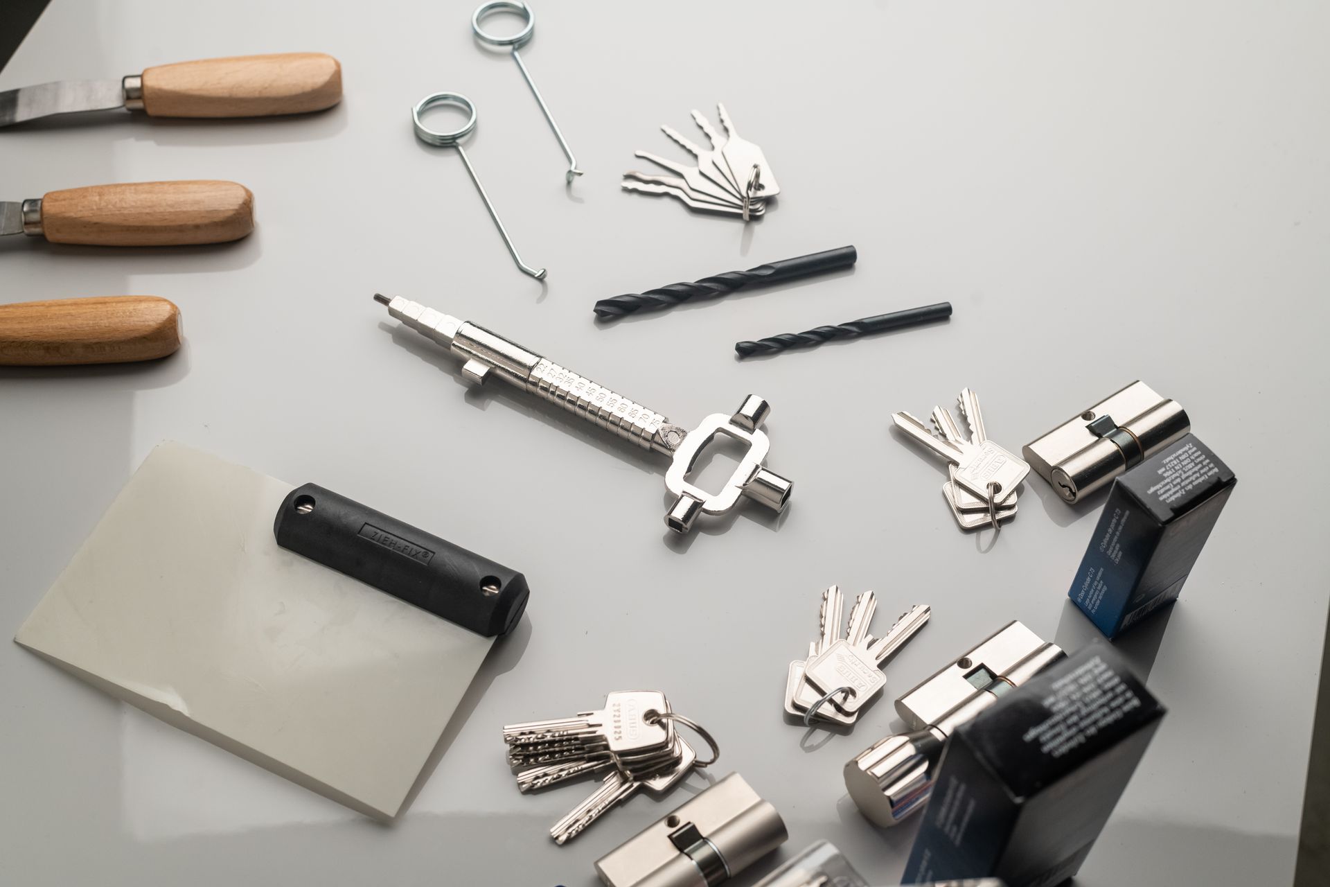 Outils de serrurier et ébauches de clés disposés sur une surface gris clair, comprenant des crochets, des clés et des forets.