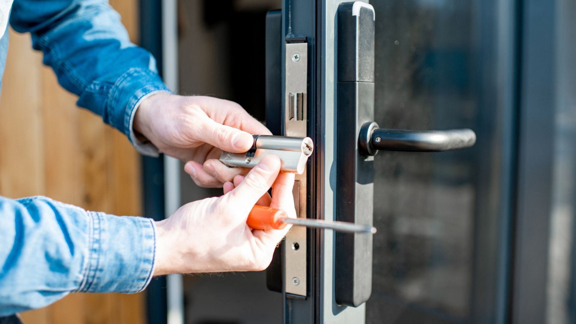 Une personne installe une serrure de porte à l'aide d'un tournevis. Gros plan sur ses mains et la quincaillerie de la porte.