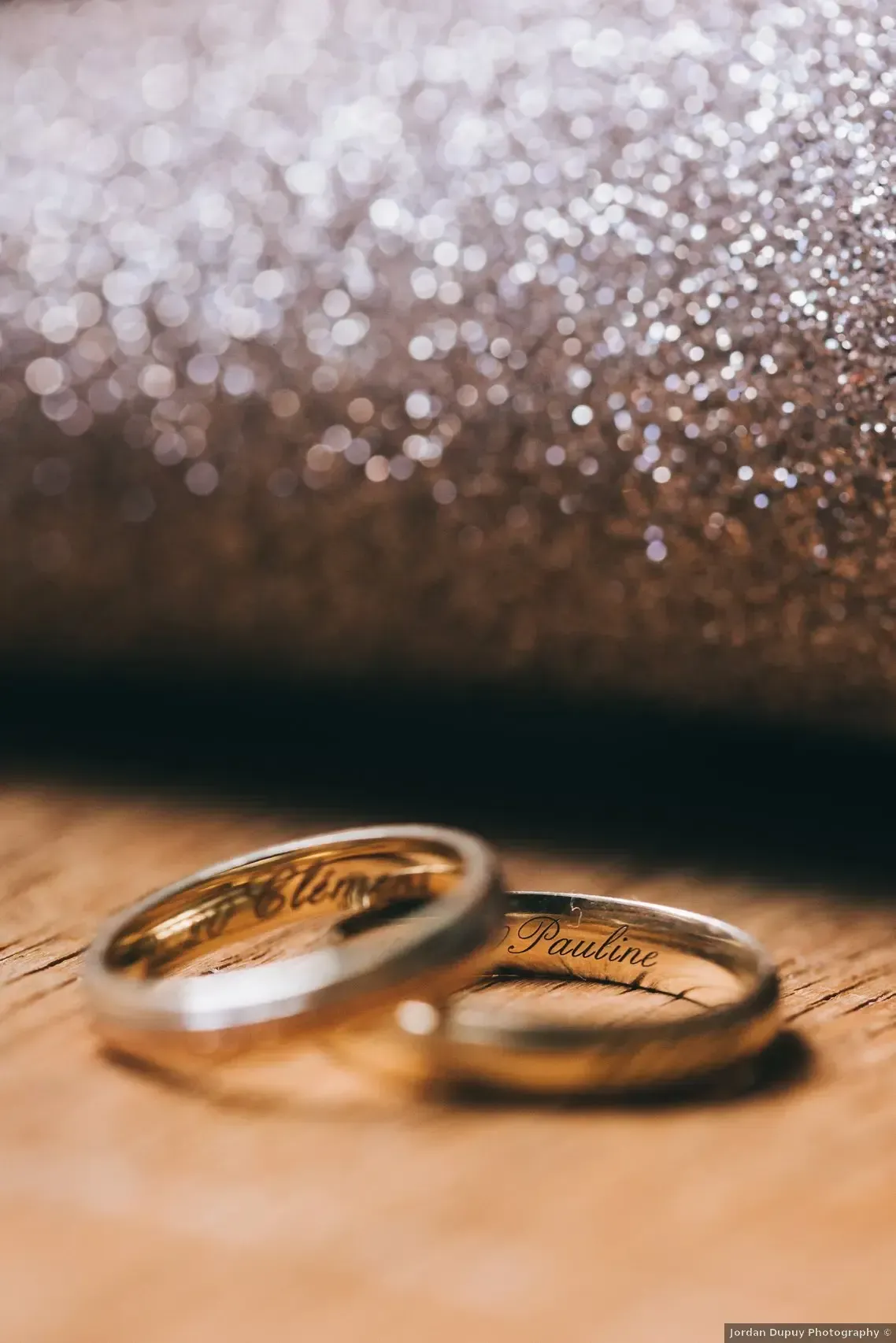 Deux alliances en or avec inscriptions gravées, sur une surface en bois, avec un fond scintillant.