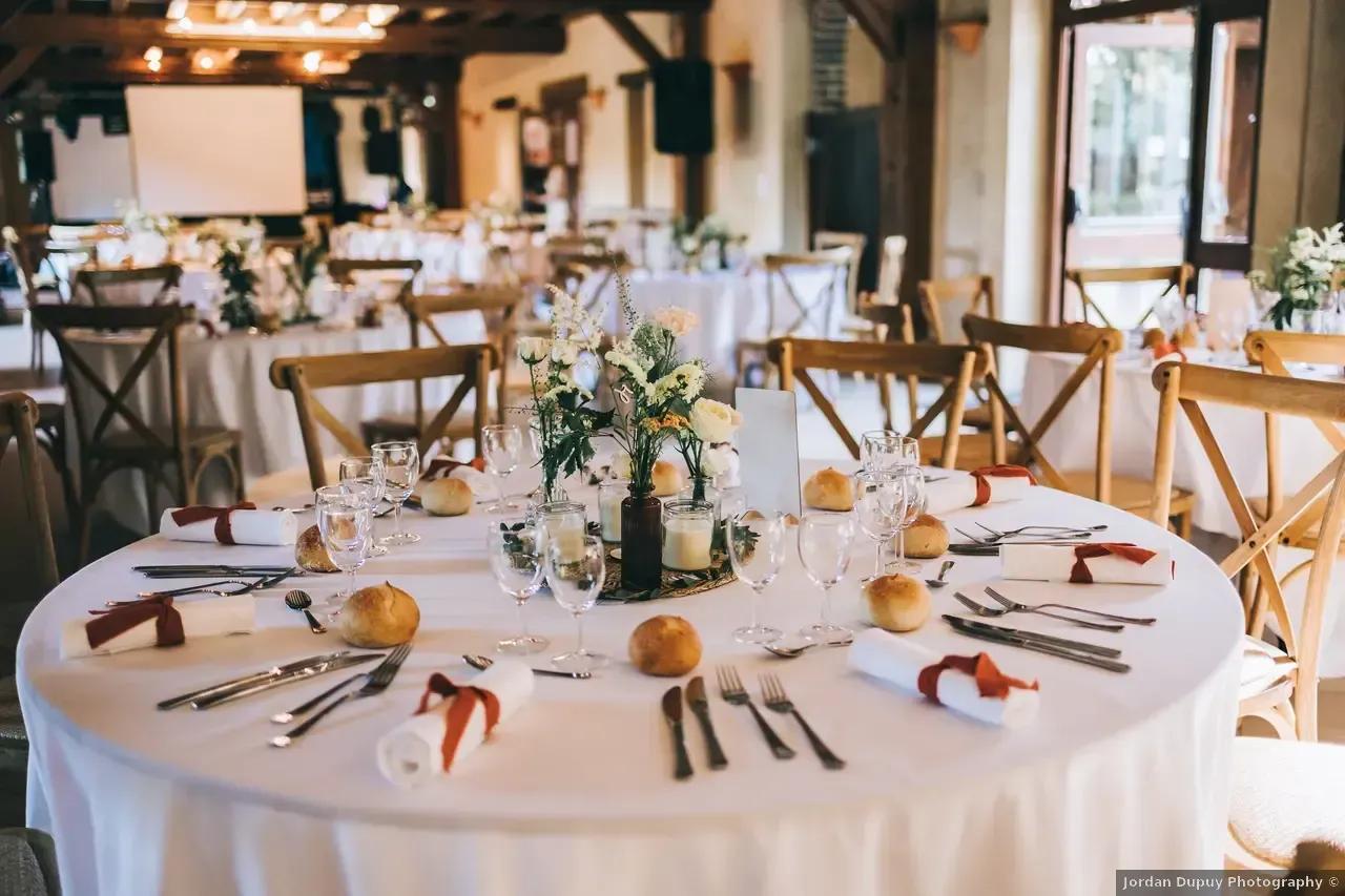 Tables rondes dressées pour une célébration dans une salle de style rustique ; nappes blanches, centres de table floraux.