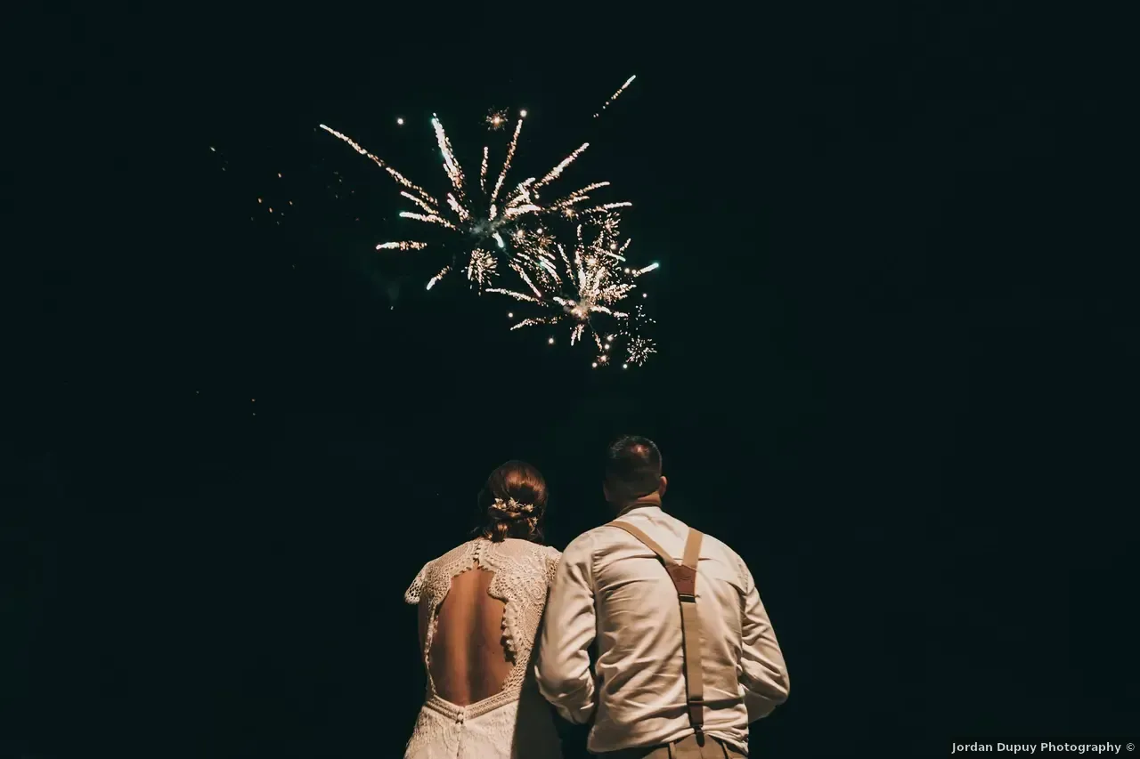 Couple regardant un feu d'artifice la nuit, vu de dos. La femme porte une robe blanche, dos nu.