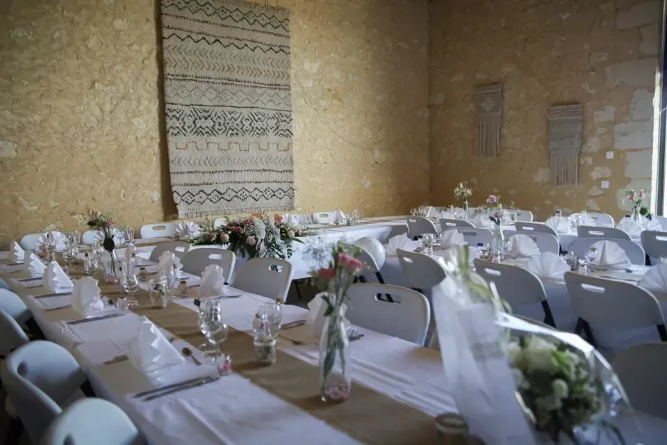De longues tables dressées pour une réception de mariage dans une pièce aux murs de pierre avec une tenture murale tissée.