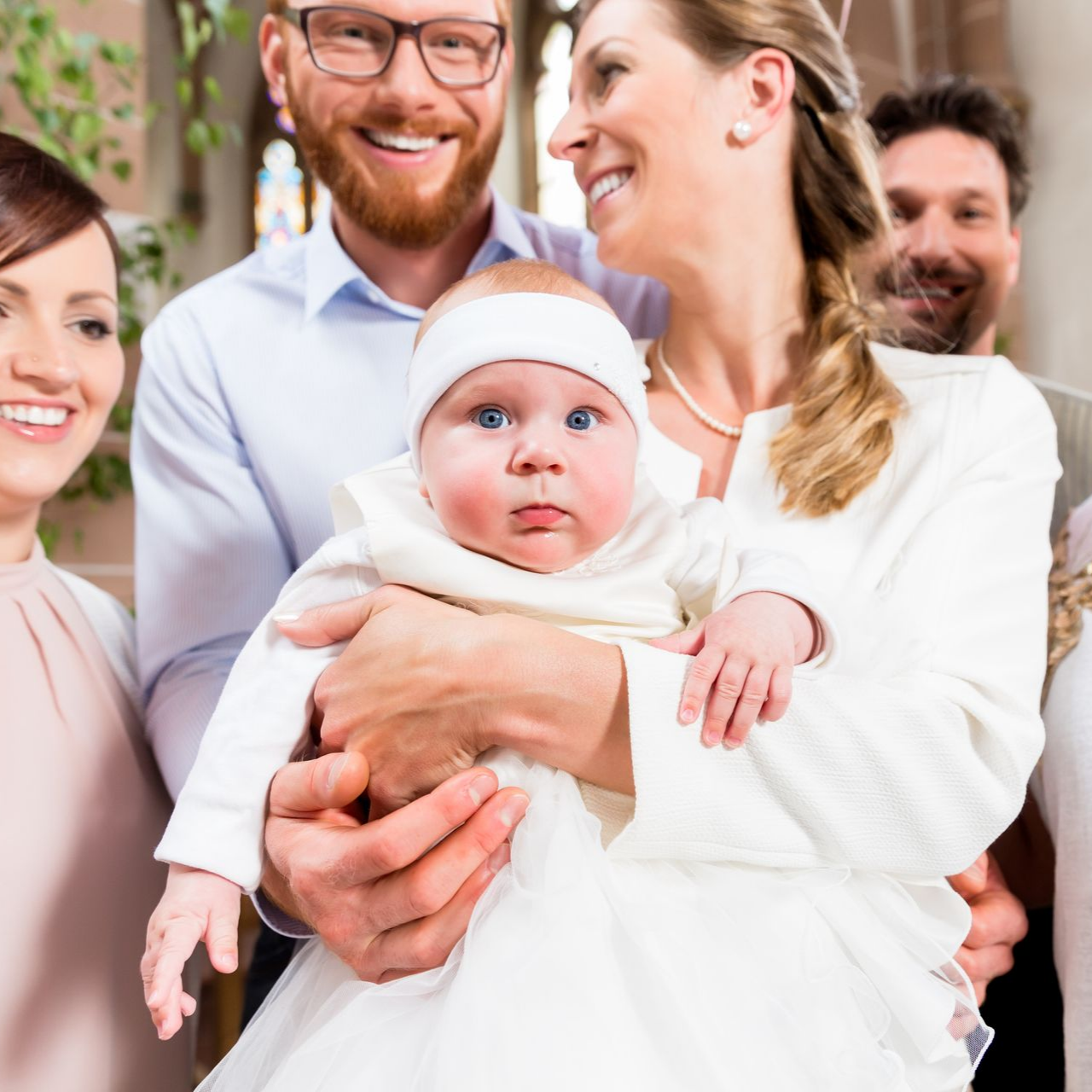 Bébé en robe blanche et bandeau tenu par une femme, entouré d'adultes souriants dans une église.