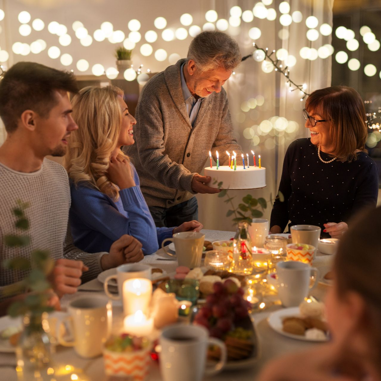 Une famille fête un anniversaire à l'intérieur. Un homme âgé tient un gâteau avec des bougies allumées.