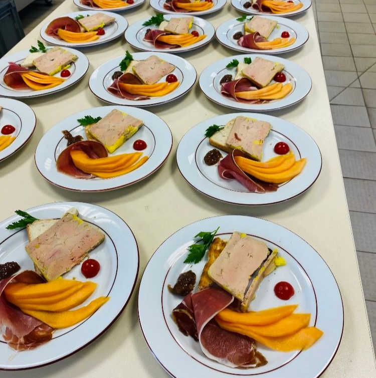 Des assiettes de nourriture, comprenant du melon, du jambon et un carré de pâté, sont disposées sur une surface blanche.