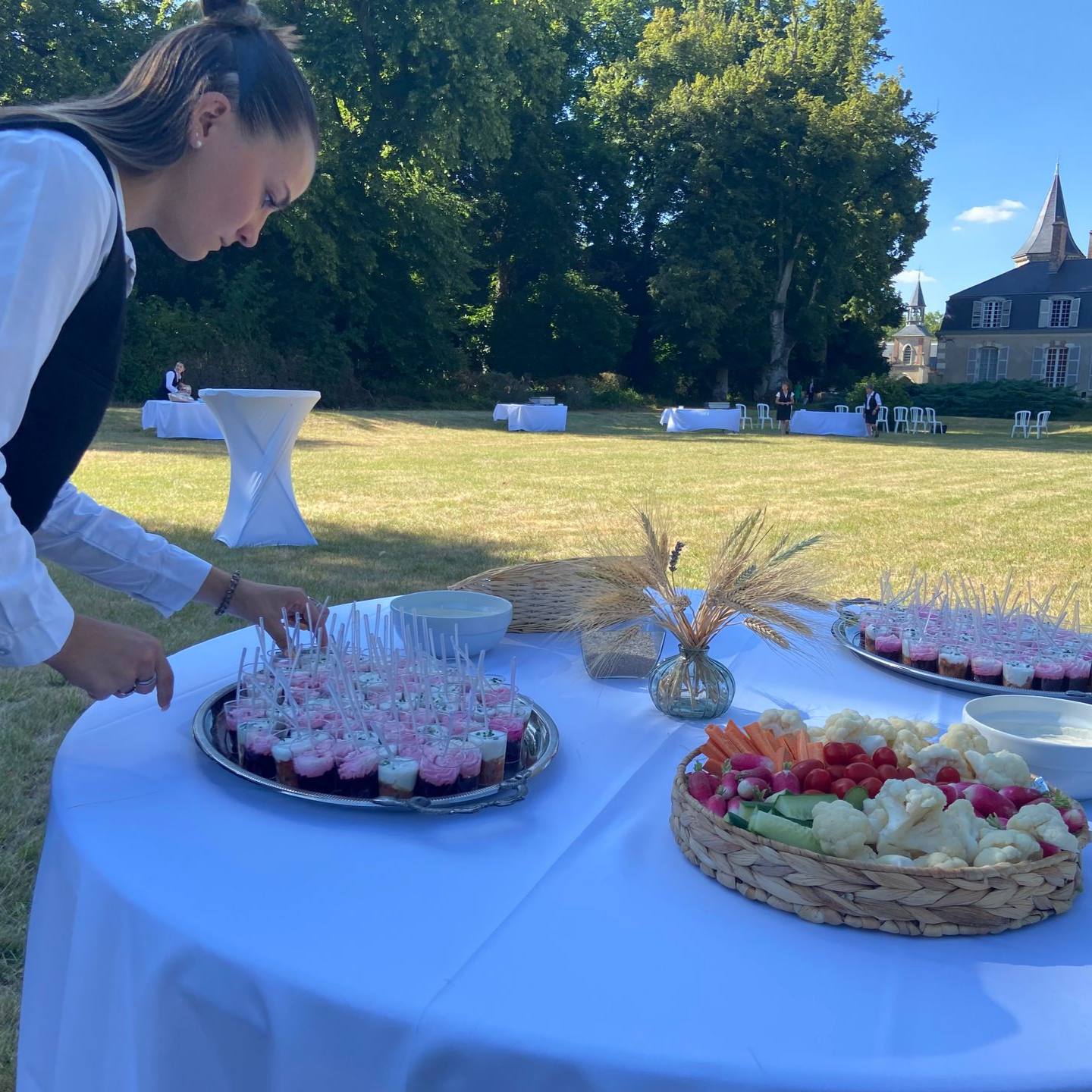 Une femme dispose de la nourriture sur une table à l'extérieur ; pelouse et bâtiment en arrière-plan.