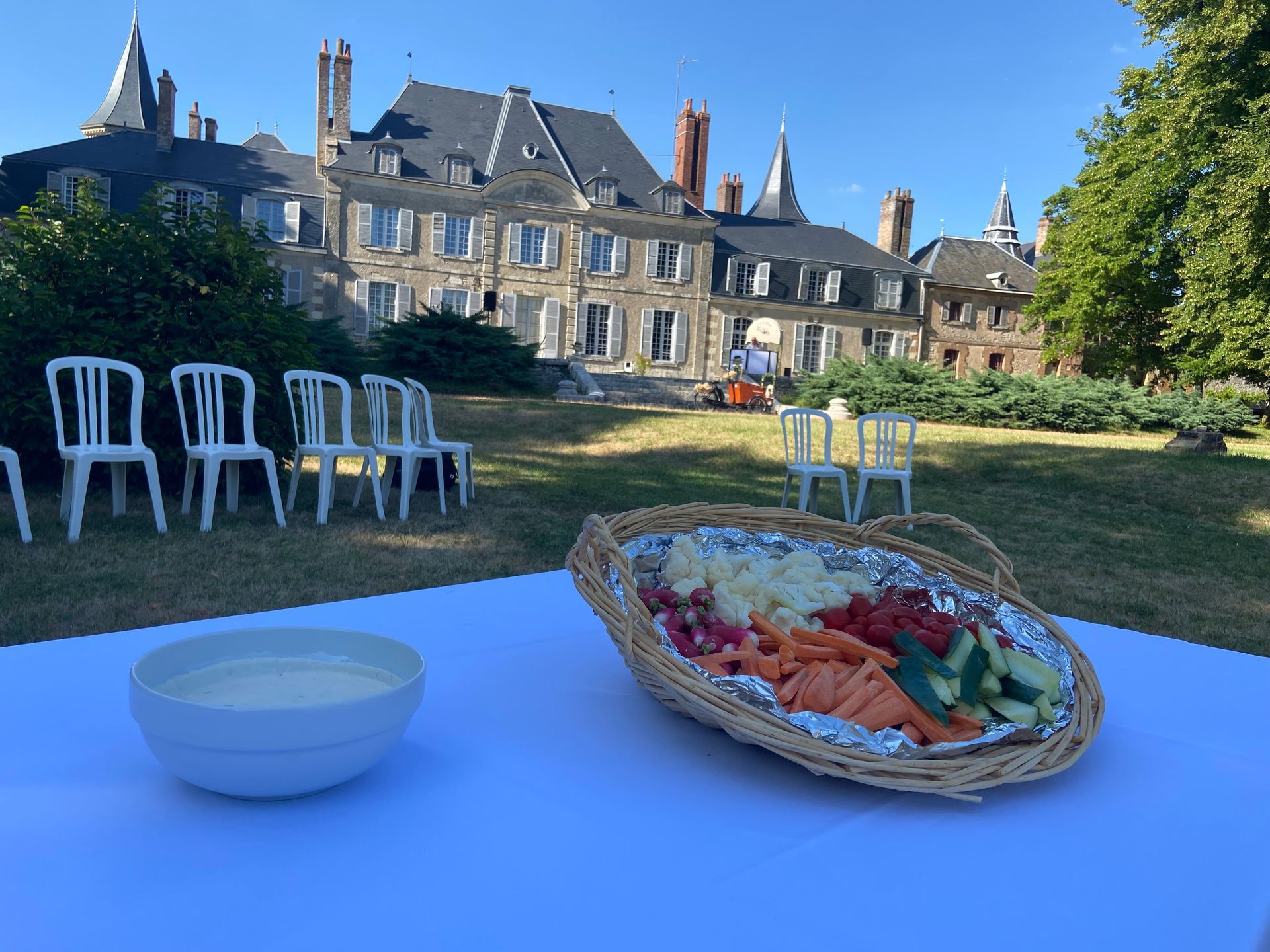 Assiette de crudités et sauce sur une table devant un grand bâtiment et une pelouse avec des chaises.
