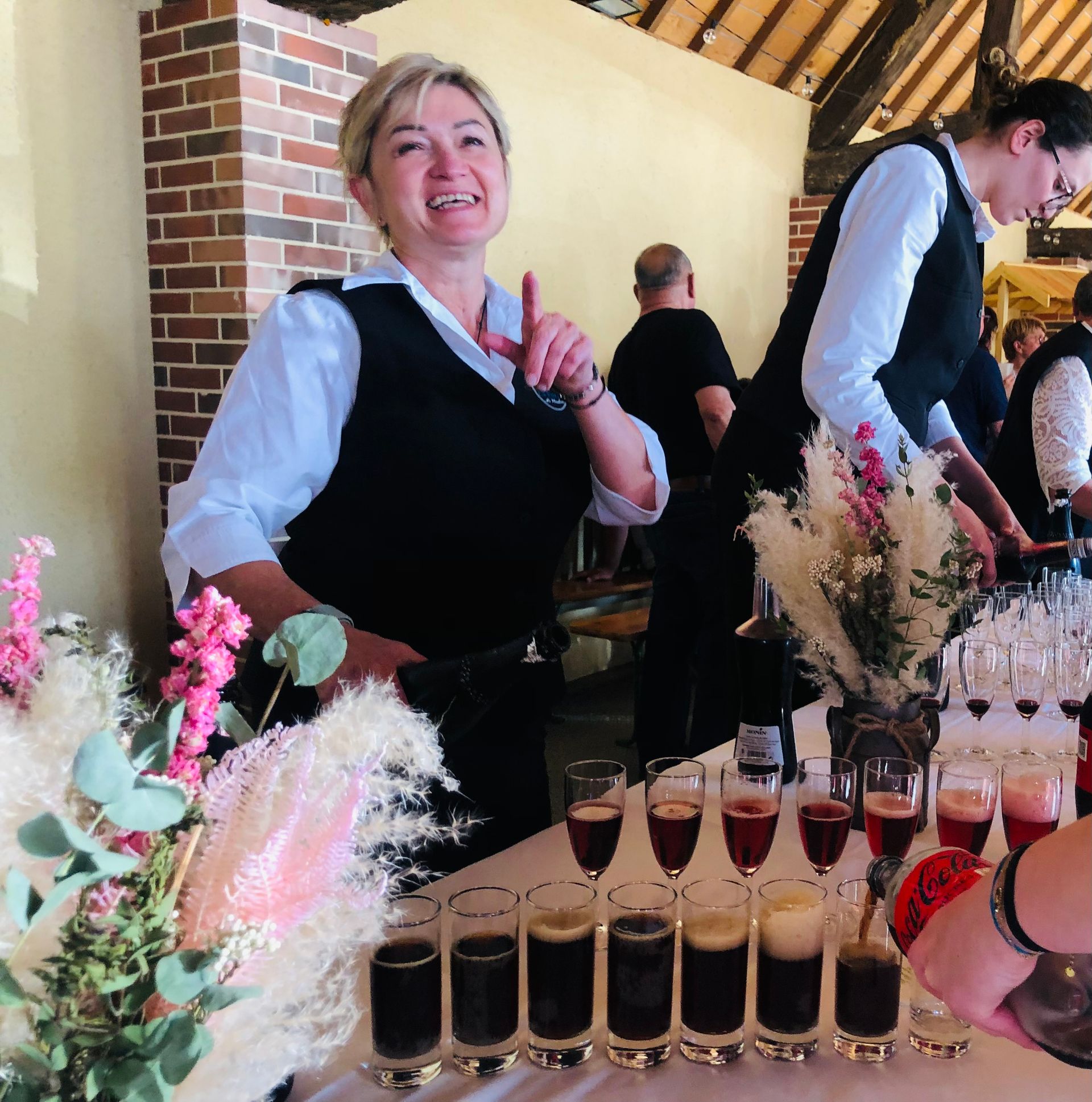 Femme derrière un bar pointant vers le haut, servant des boissons noires lors d'un événement, avec des décorations florales.