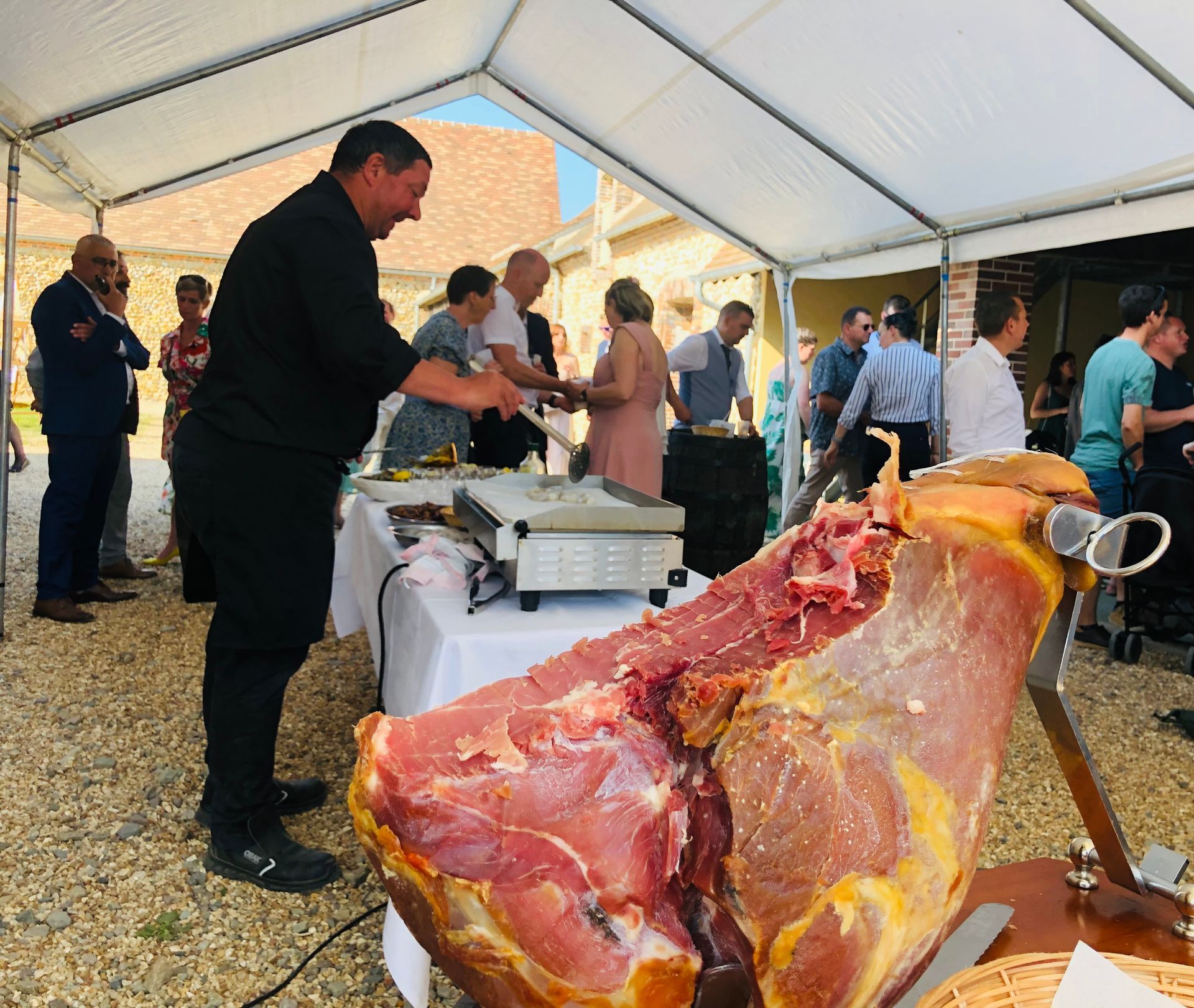 Un homme découpe du jambon lors d'un événement avec traiteur sous une tente, avec des invités attendant à proximité.