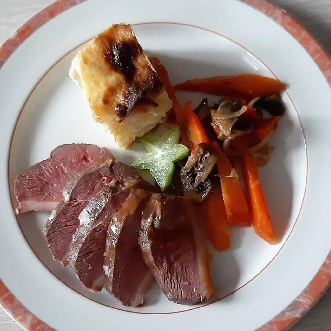 Magret de canard tranché, légumes rôtis et gratin de pommes de terre sur une assiette blanche.