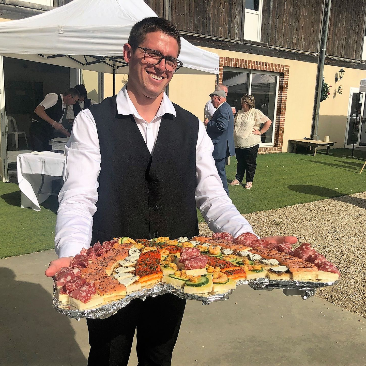 Un homme souriant tient un plateau d'amuse-gueules dehors. D'autres personnes et une tente sont visibles à l'arrière-plan.