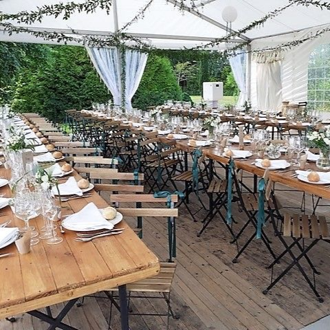 De longues tables en bois dressées pour un repas sous une tente blanche, décorée de verdure et de rideaux, en extérieur.