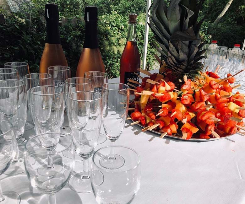 Table dressée pour la fête : bouteilles de champagne, brochettes de fruits et verres vides sur une nappe blanche.